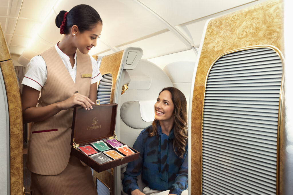Flight attendant smiling as she presents a selection of teas to a seated passenger in a luxurious first-class cabin. The atmosphere is warm and elegant.