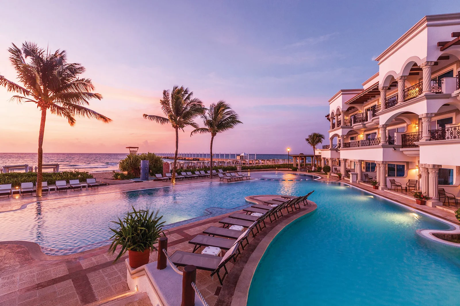 Image shows the Hilton Playa del Carmen Sunset Pool at sunset with palm trees, lights and seats overlooking the view
