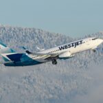 A WestJet airplane climbing through the sky, with snowy mountains in the background, showcasing a clear blue atmosphere.