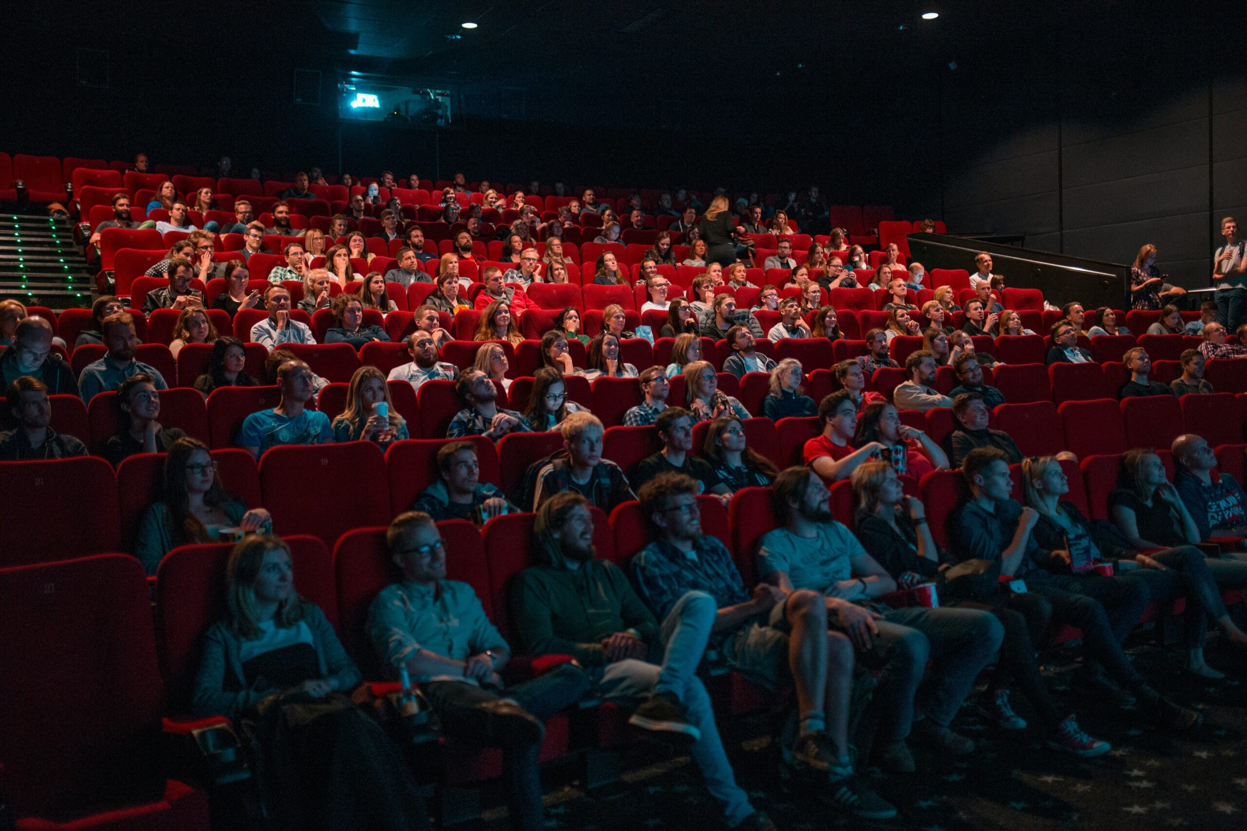 Audience in a dimly lit theatre sits in rows of red seats, focused on a screen. The atmosphere is engaged and anticipatory, with varied expressions.
