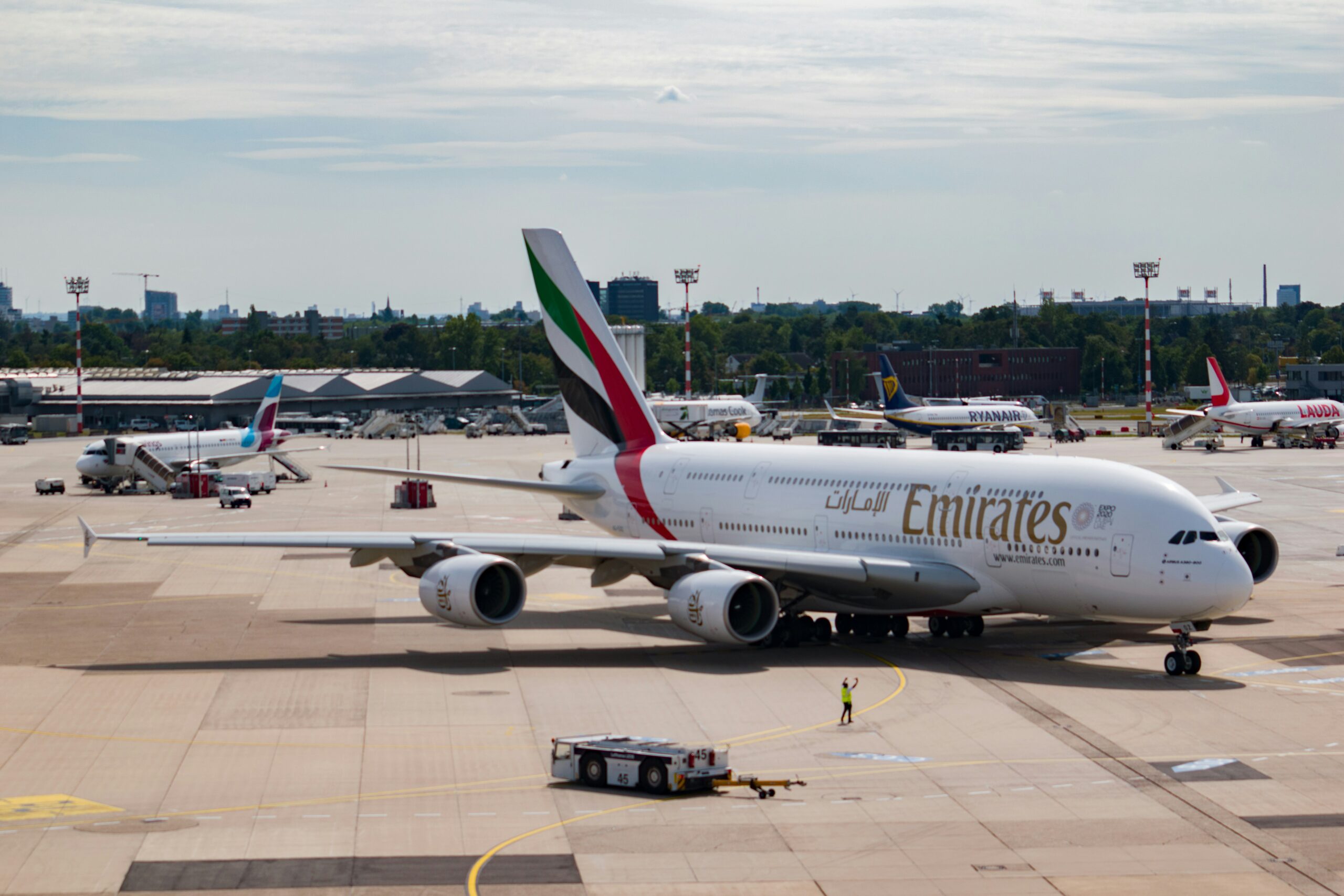 An Emirates Airbus A380 is parked on an airport tarmac with various other aircraft and ground vehicles visible in the background.