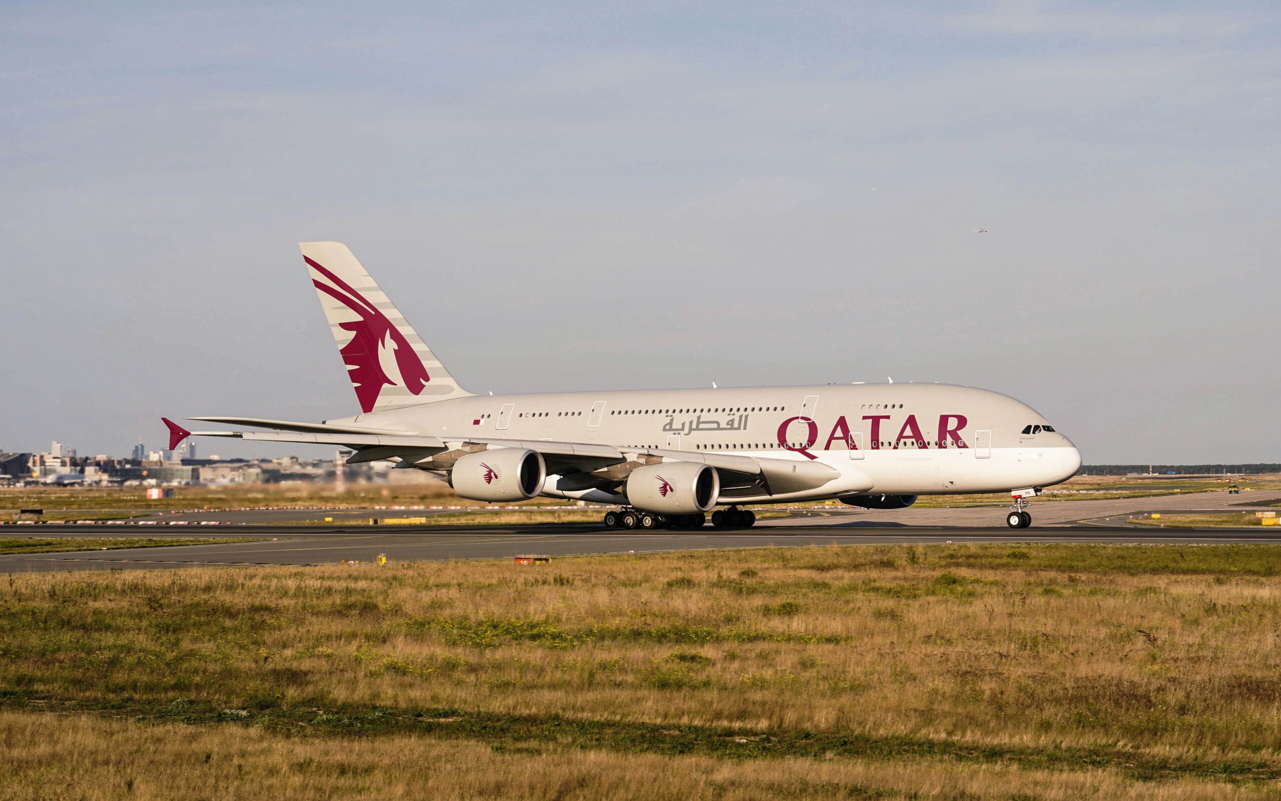 Large Qatar Airways aircraft with maroon logo on the tail, taxiing on a runway. The background shows a sunny sky and distant cityscape.
