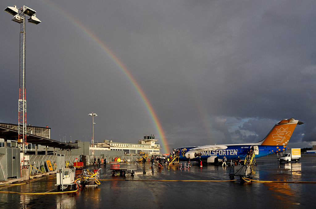 An image of Stockholm Airport in the rain. There is a double rainbow behind the plane and the airport on the tarmac.