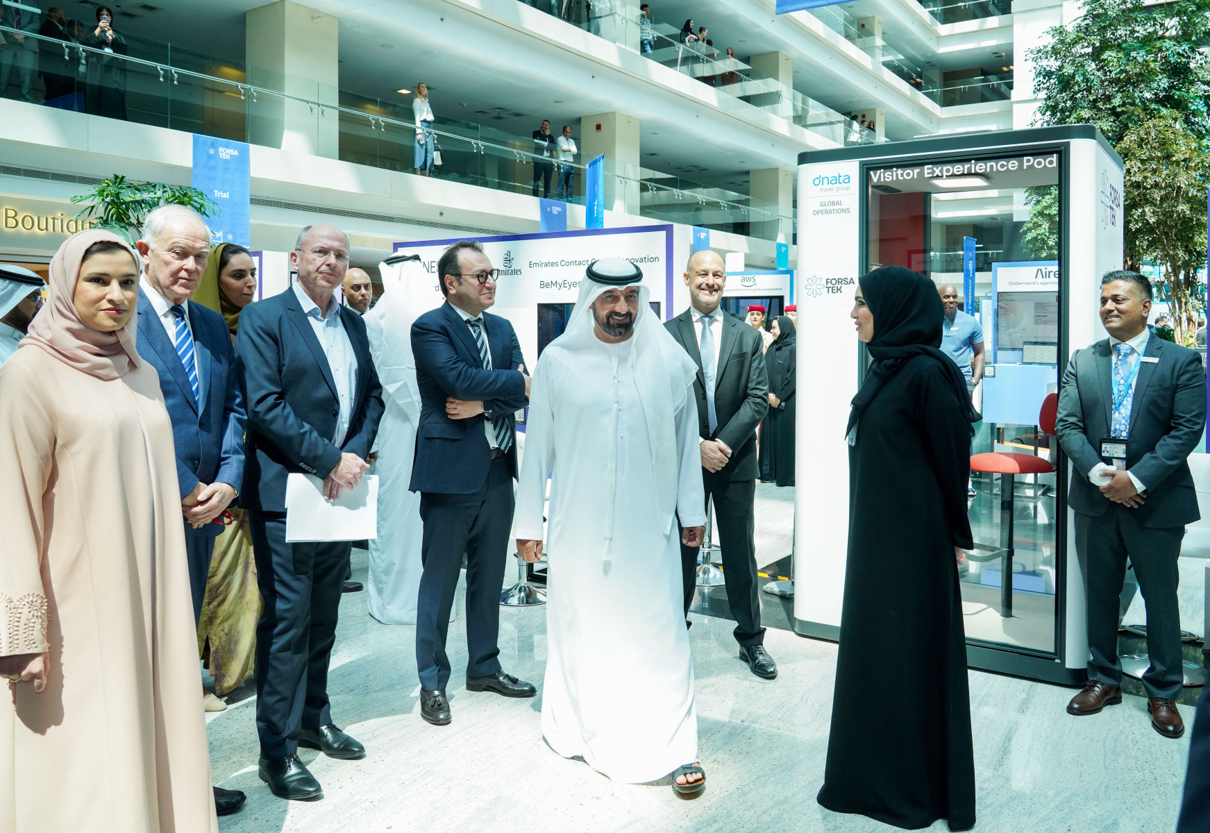 A group of people, including men in suits and traditional Middle Eastern attire, stand in a modern exhibition space near a “Visitor Experience Pod” display.