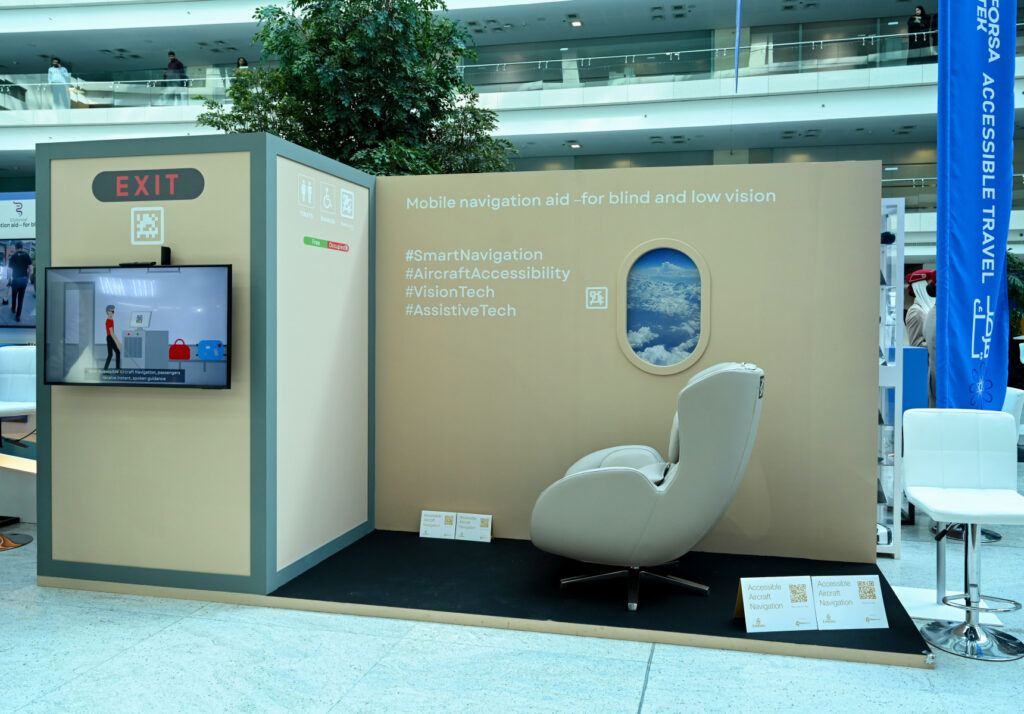 Exhibit booth displays a navigation aid for blind and low vision users, with a chair, a screen, informational signs, and accessibility hashtags on a beige wall.