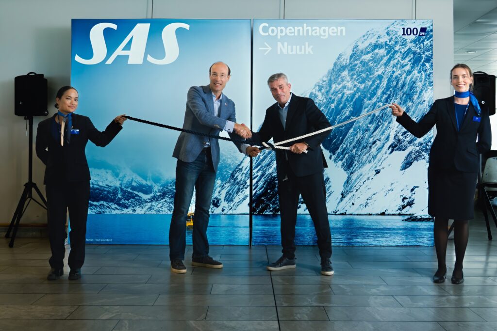 Four people in standing, two of which are in the middle and holding a half black and half white rope which is knotted together. They are shaking hands. Photo represents new flight route from Copenhagen, Denmark to Nuuk, Greenland.
