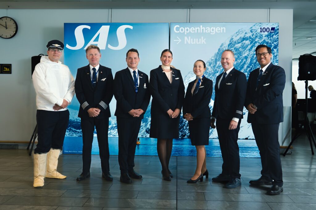 Cabin crew members are smiling and standing in front of a blue background with a mountain that says SAS and Copenhagen-Nuuk representing the new flight route.