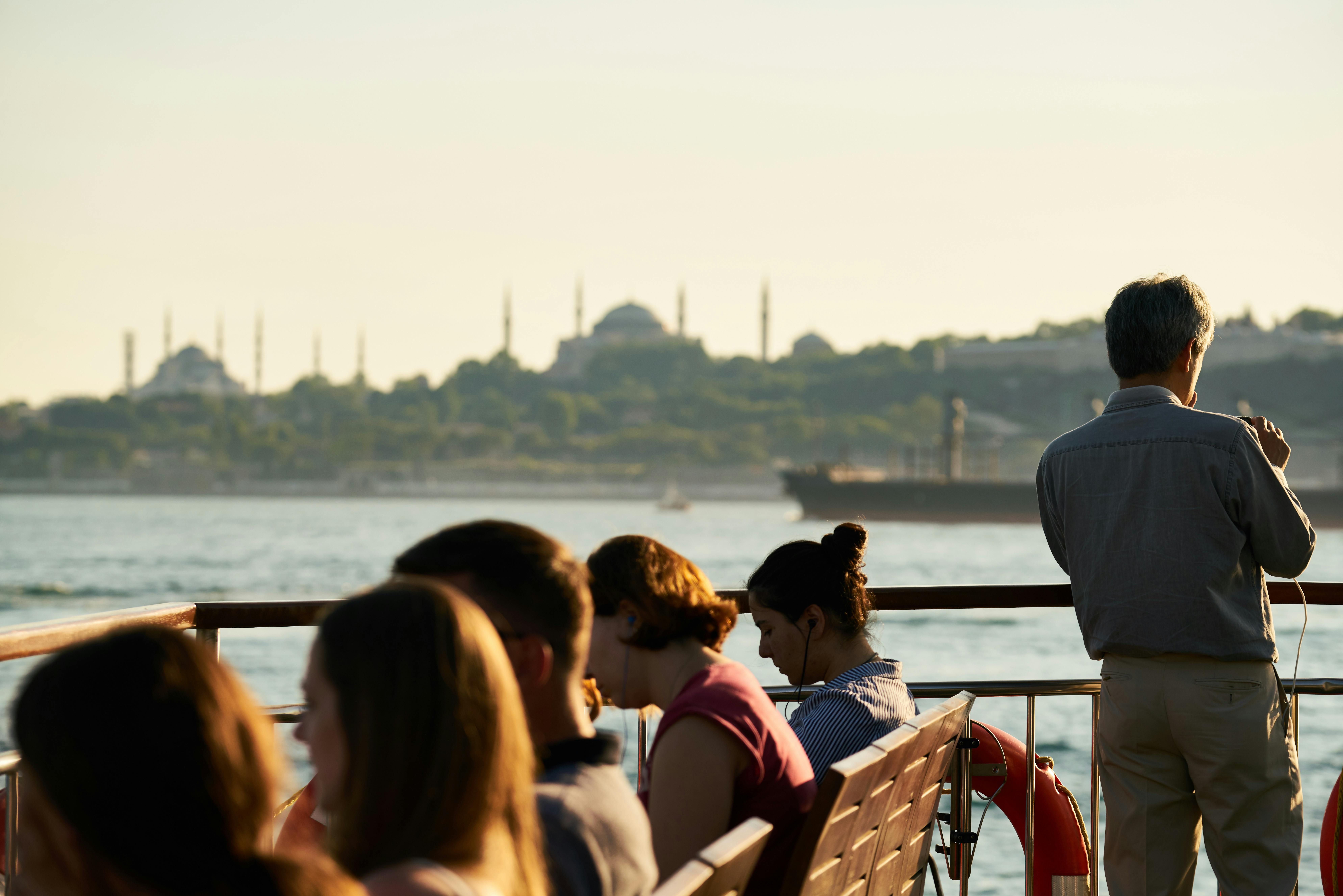 People sitting on a boat and a man standing by the railings with a panoramic view of Istanbul in the background