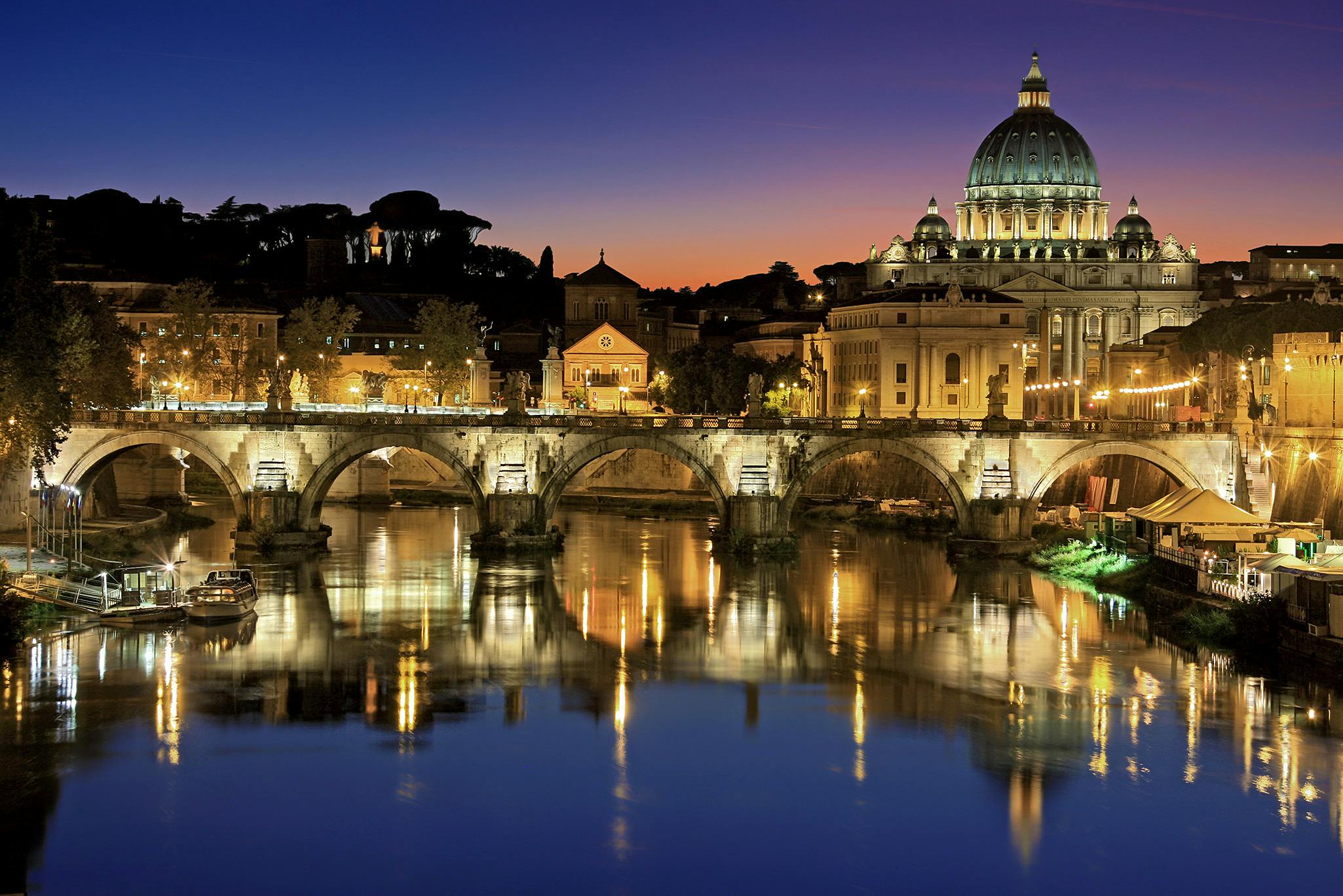 A view of Vatican City at dusk. St Peter's Basilica is illuminated with a bridge in front of it and a reflection in the water below.