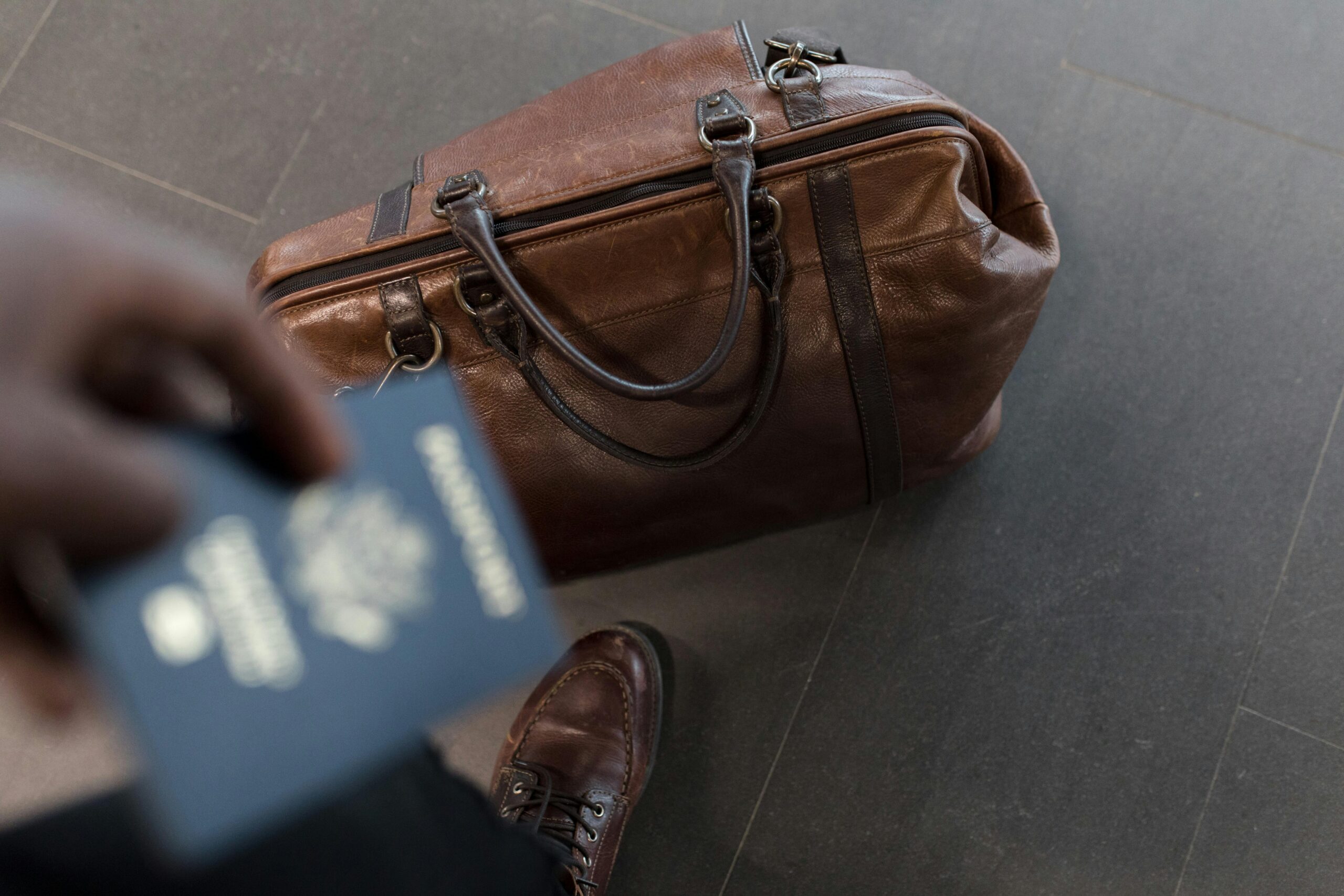 A men holding a passport in one hand and a brown leather duffel bag on the ground