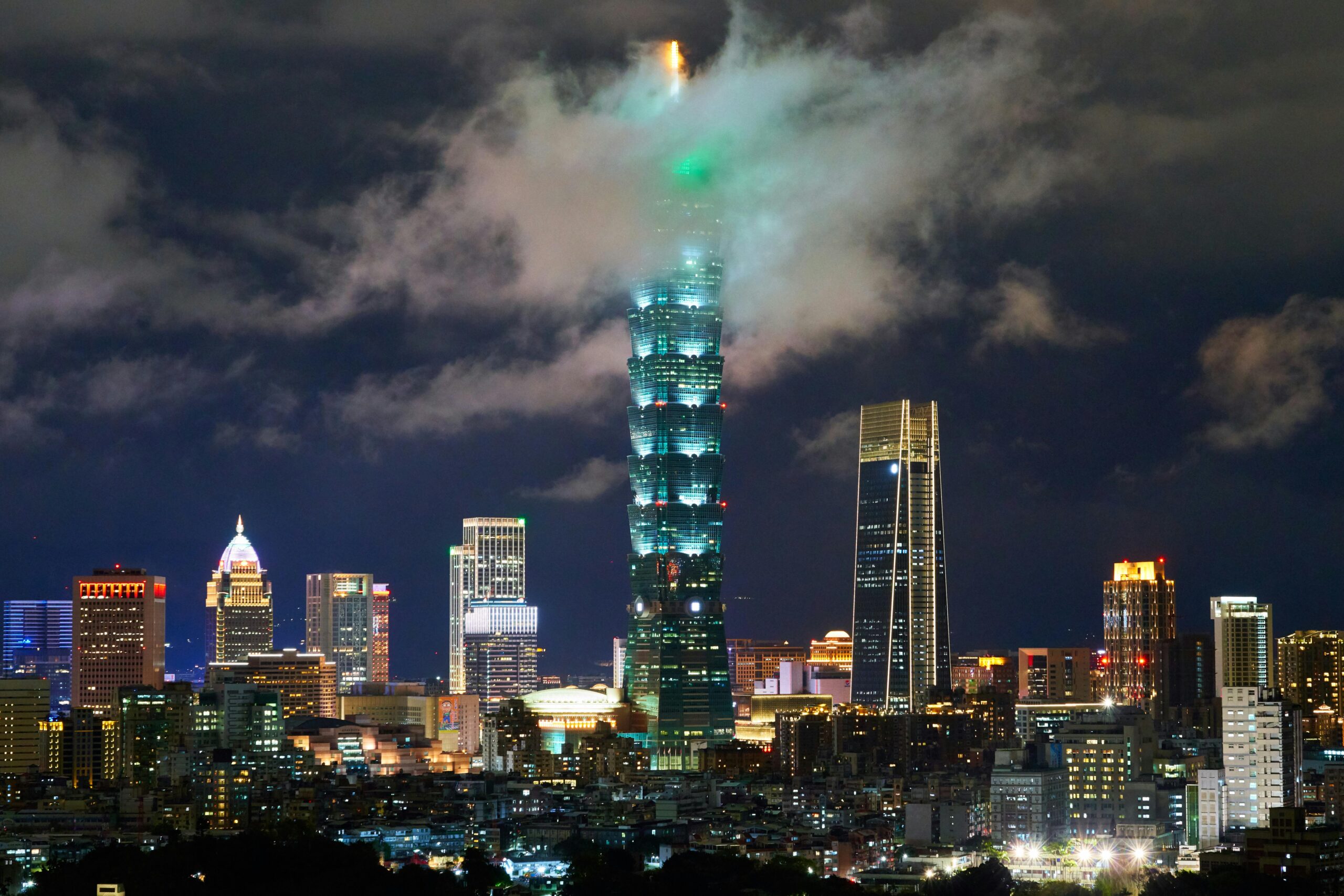 An image of Taipei's skyline at night with a tall skyscraper in the centre poking through the clouds