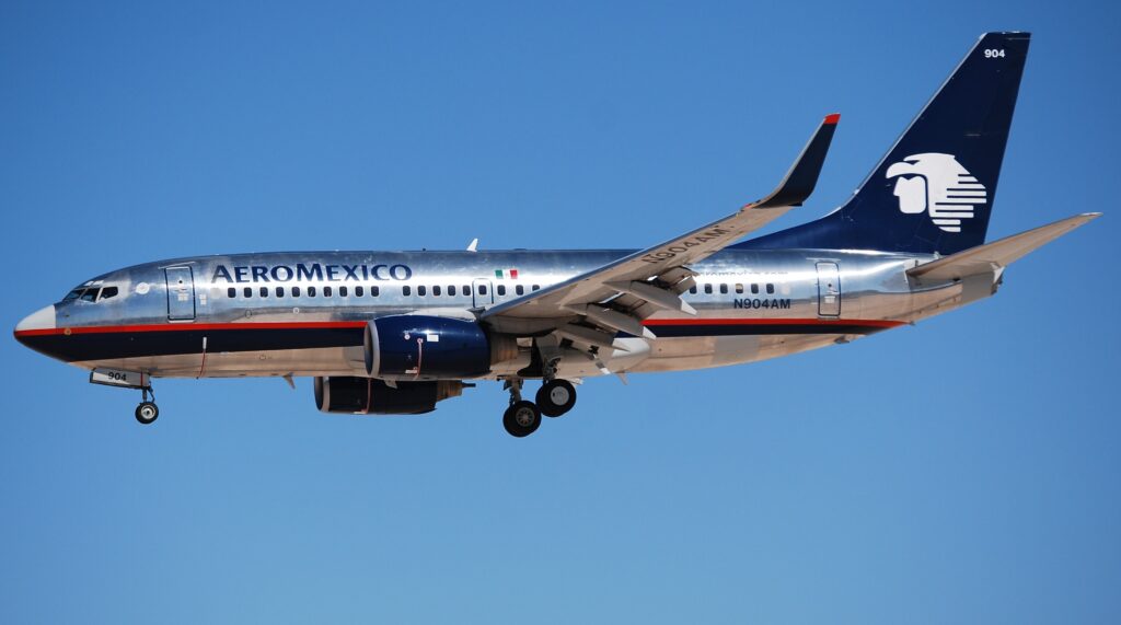 Silver Aeromexico jet with a blue tail fin. Aeromexico is written in bold capitals. The background is a plain blue sky.