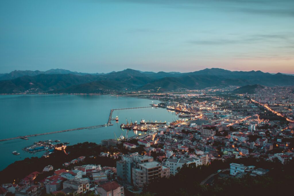 Evening scene of Béjaïa, Algeria, with the cityscape glowing softly under a dusky sky, reflecting on the water.