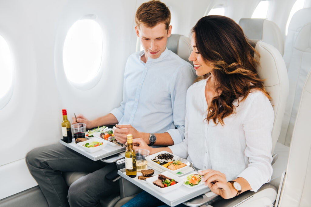 Two people dining on an aeroplane tray, featuring their meal and the aeroplane's interior setting around them.