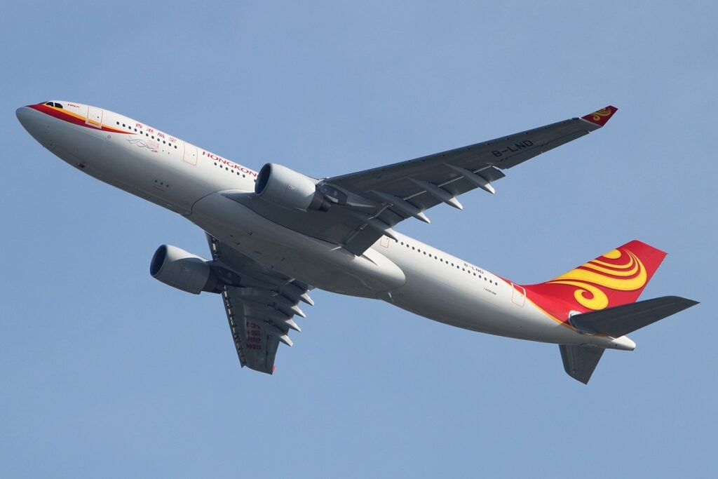 A Hong Kong Airlines jet soaring through a clear blue sky, showcasing its wings and tail against the backdrop of clouds.