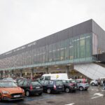 Exterior view of Il Caravaggio International Airport (Bergamo Orio al Serio Airport) with parked cars and passengers entering the terminal on a cloudy day.