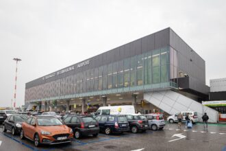 Exterior view of Il Caravaggio International Airport (Bergamo Orio al Serio Airport) with parked cars and passengers entering the terminal on a cloudy day.