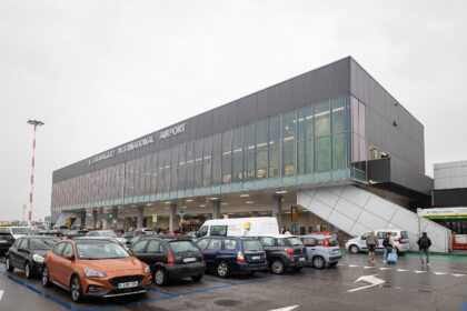 Exterior view of Il Caravaggio International Airport (Bergamo Orio al Serio Airport) with parked cars and passengers entering the terminal on a cloudy day.