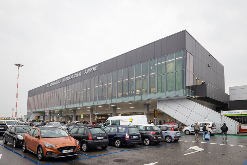 Exterior view of Il Caravaggio International Airport (Bergamo Orio al Serio Airport) with parked cars and passengers entering the terminal on a cloudy day.