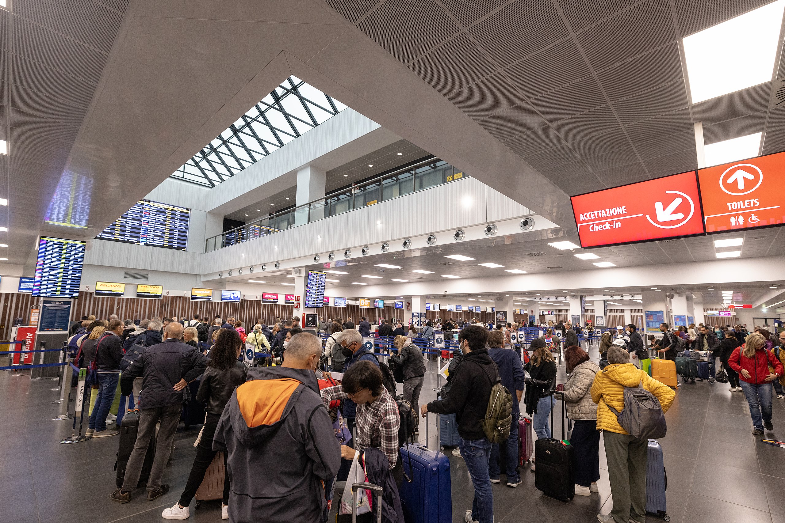 Busy check-in area inside Bergamo Airport with passengers queuing, digital flight information displays, and directional signs in Italian.