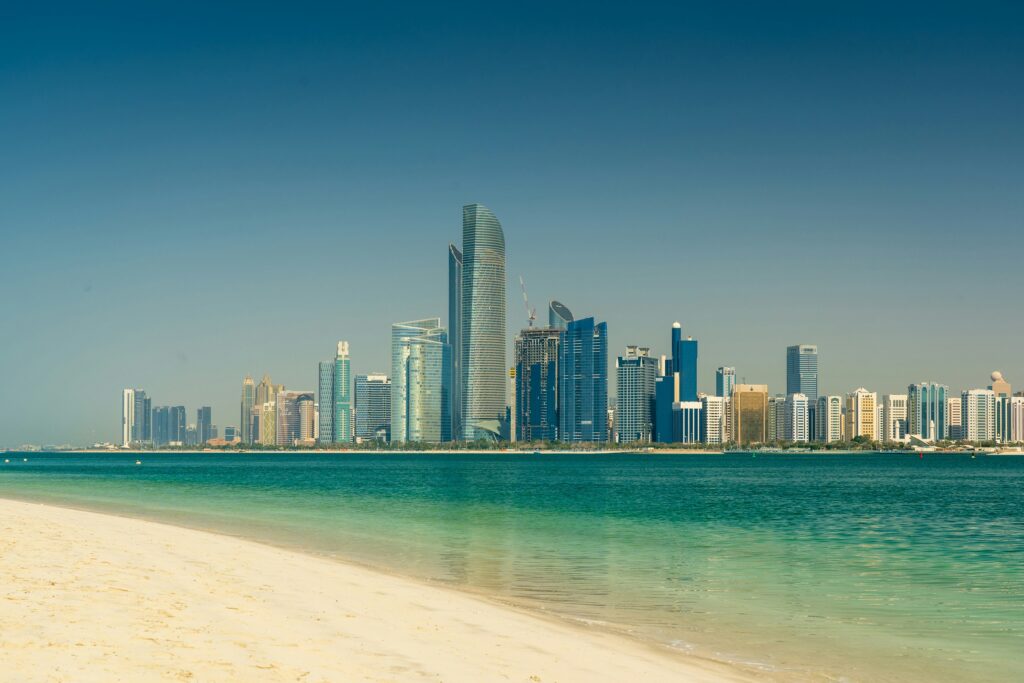 Wide view of a serene beach with turquoise water in the foreground, set against a backdrop of a modern skyline under a clear blue sky.