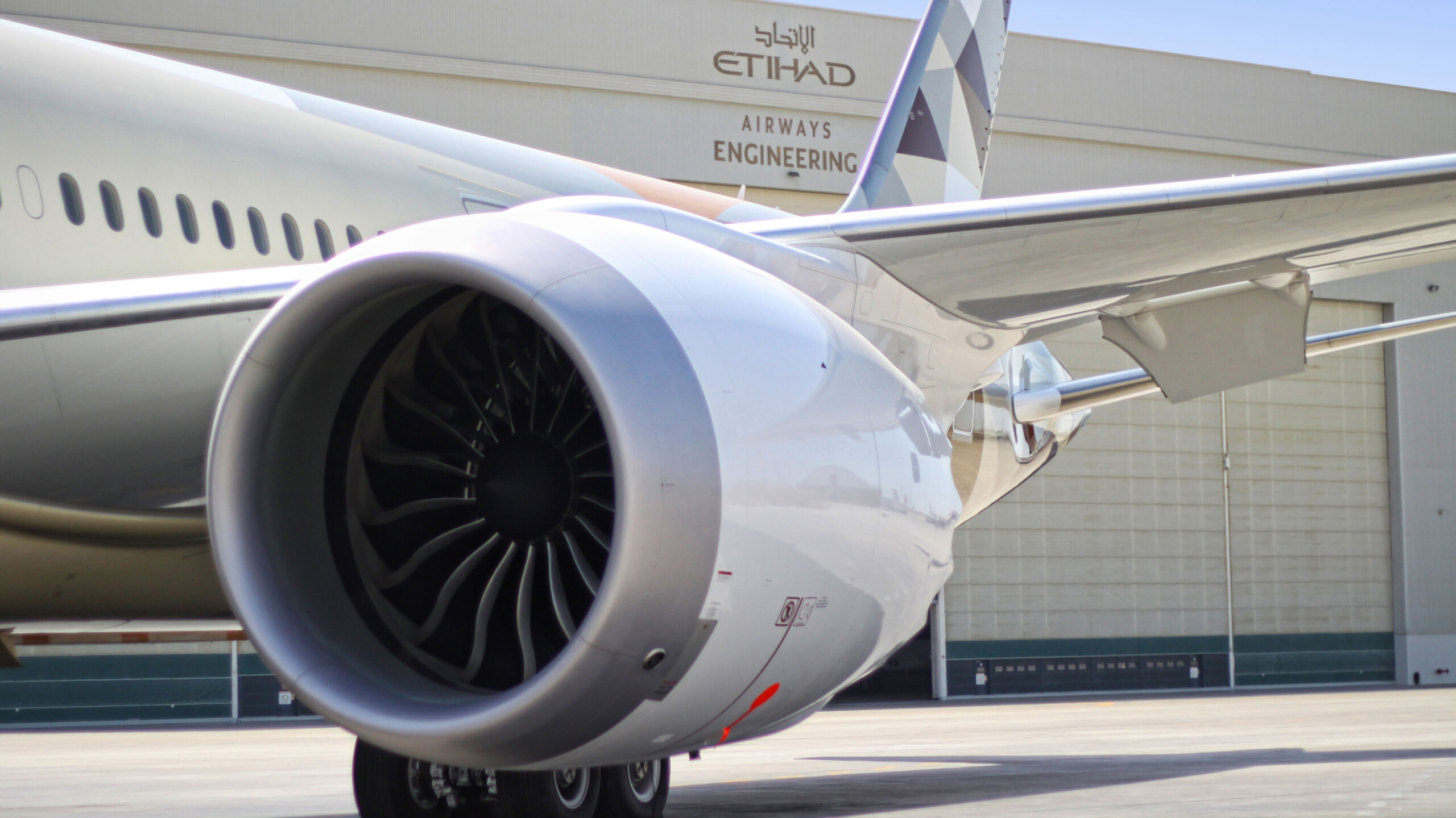 Close-up of a jet engine on a parked airplane near an Etihad Airways Engineering hangar. The sleek metal and bold lines convey precision and power.