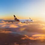 An Aeromexico Connect Embraer E190 aircraft flying above the clouds during sunset, with golden light illuminating the sky and cloudscape.