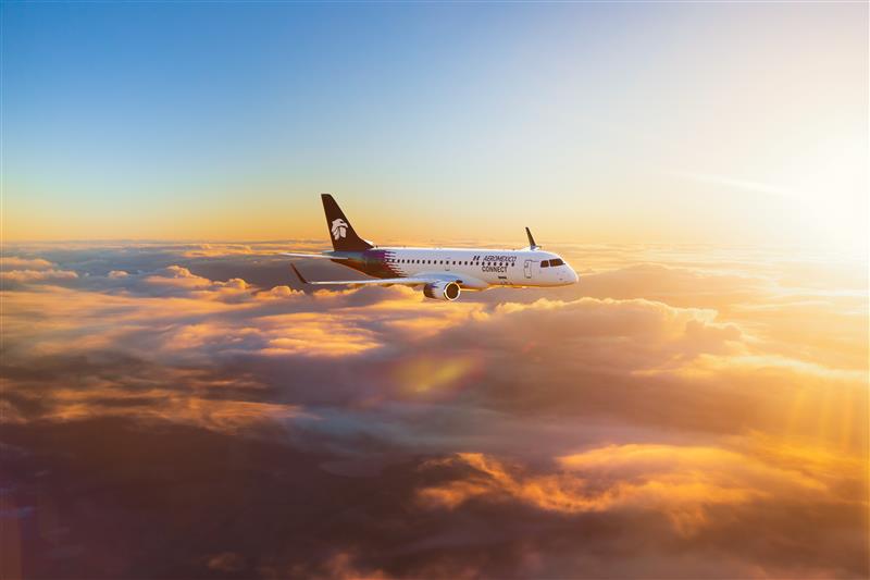 An Aeromexico Connect Embraer E190 aircraft flying above the clouds during sunset, with golden light illuminating the sky and cloudscape.