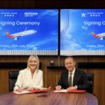 The representatives were attending the signing ceremony. Two individuals signing documents at a table, with a large screen displaying information displaying the aeroplane with the logo of Hong Kong Airlines in the background.
