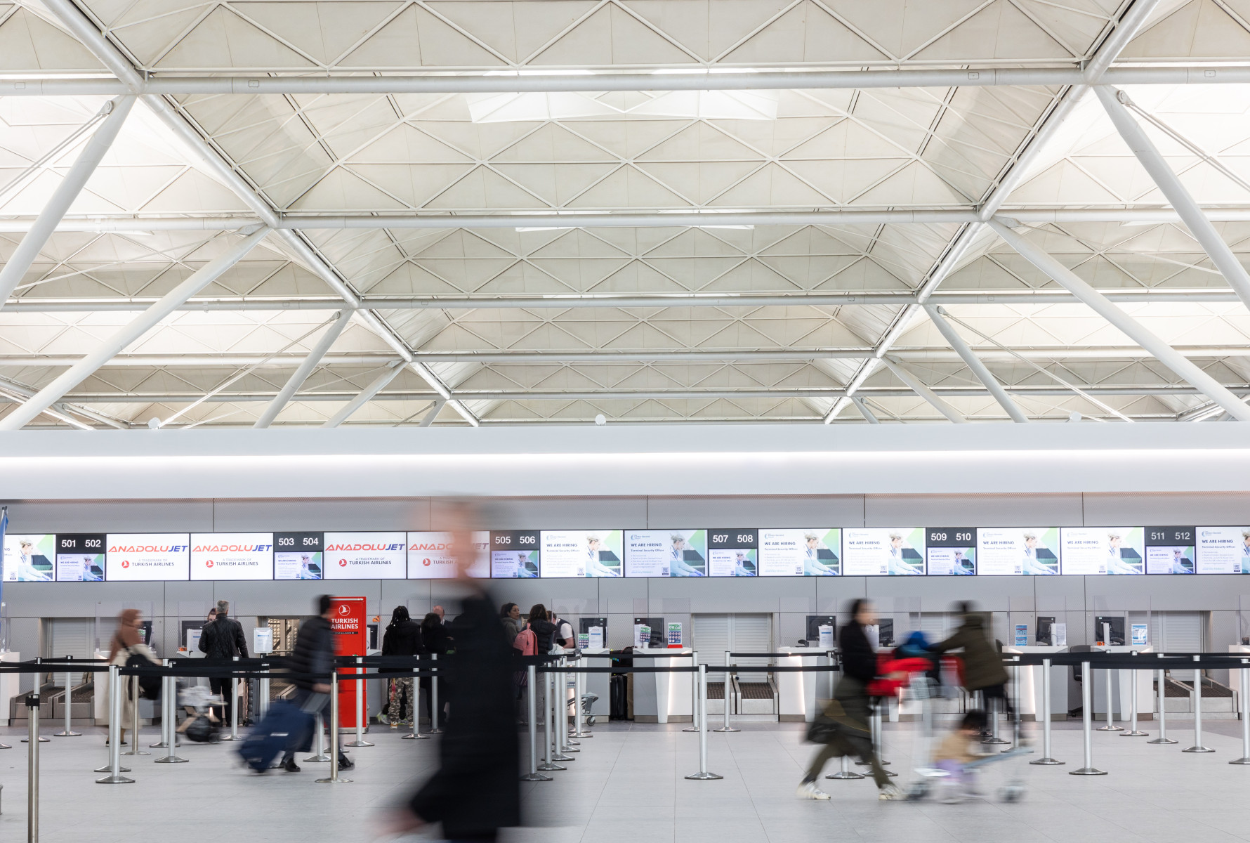 Interior of an airport check-in area with digital screens displaying flight information, counter desks, and passengers in motion.