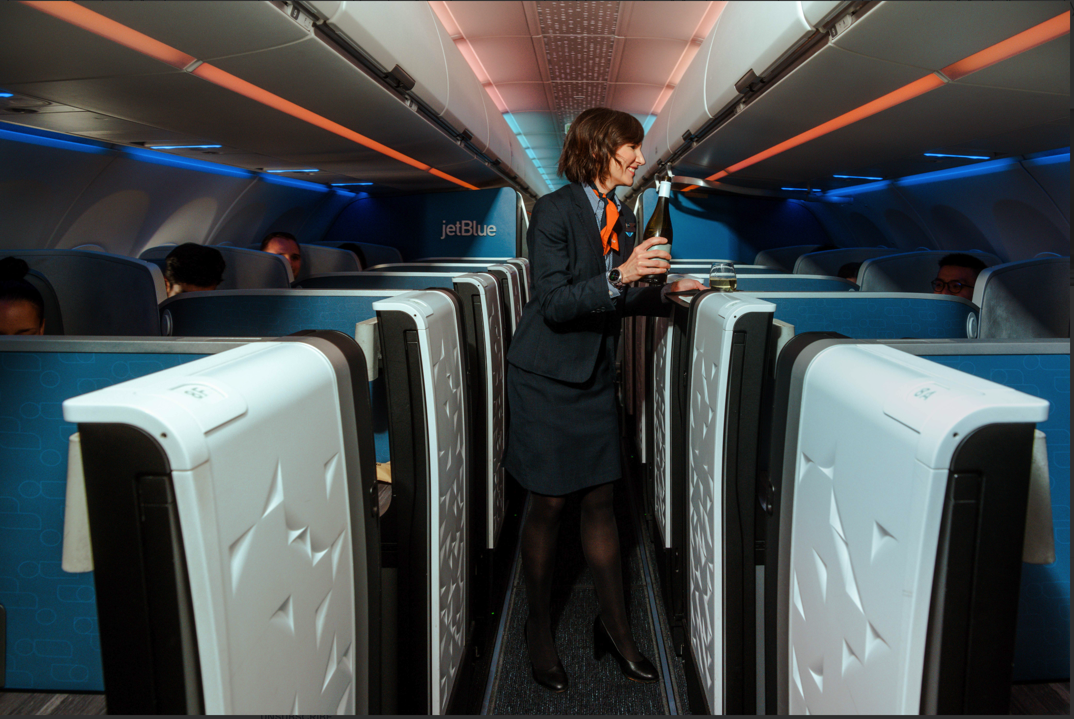 JetBlue flight attendant in navy uniform and orange scarf serving champagne in the Mint business class cabin, with private suites, mood lighting, and seated passengers.