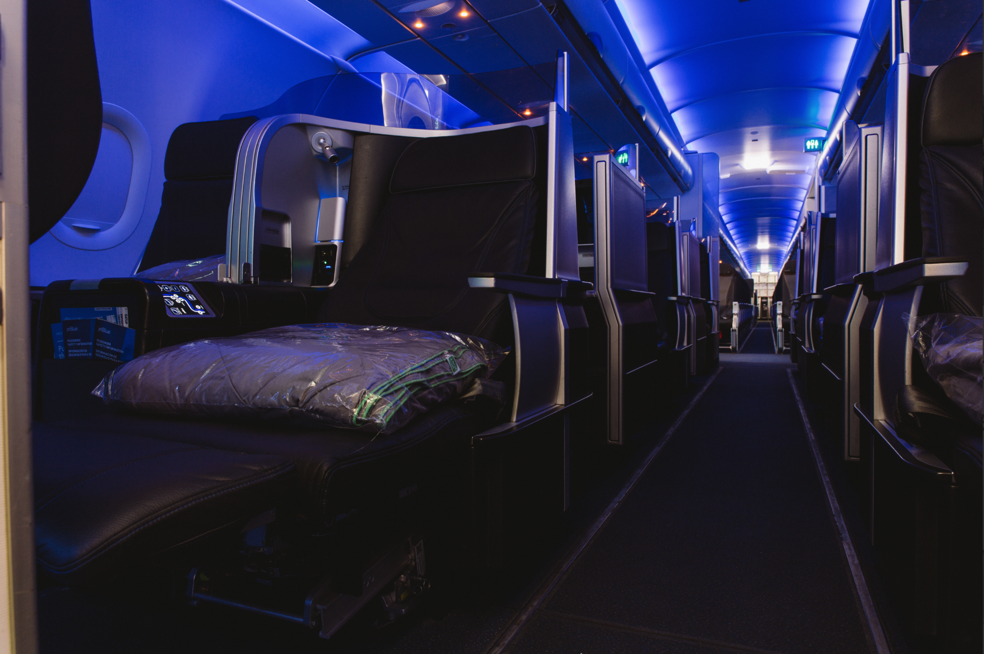 JetBlue Mint business class cabin featuring lie-flat seats with personal entertainment screens, pillows and blankets, under blue ambient lighting in a modern aircraft interior.