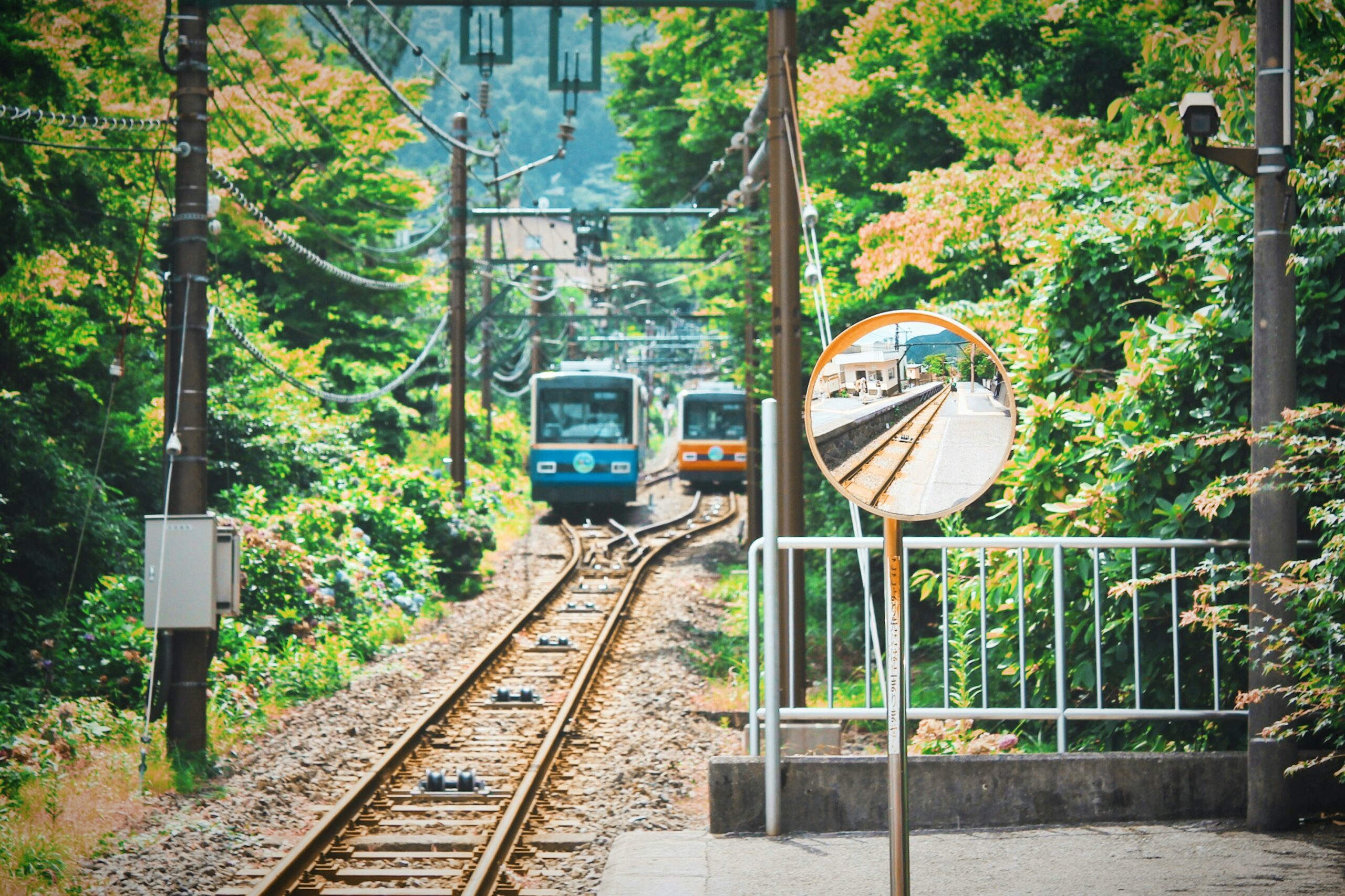 A vibrant train station surrounded by lush greenery, featuring a safety mirror reflecting the platform and train tracks in Japan.