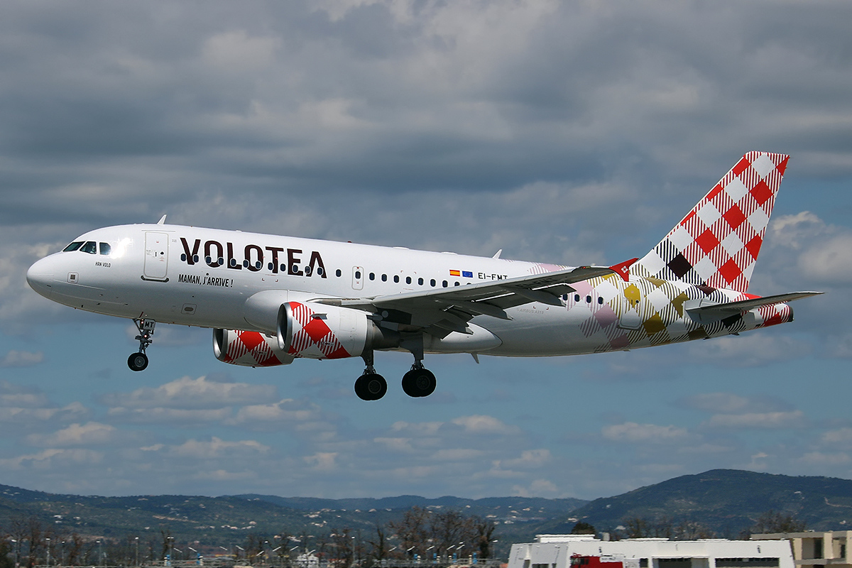 Volotea Airbus A319 aircraft in mid-flight with distinctive red checkered tail livery, flying against a partly cloudy sky.