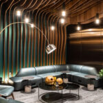 modern airport lounge interior with curved wood and teal wall paneling, soft leather seating, round black coffee tables, ambient pendant lighting, and a large arched floor lamp.