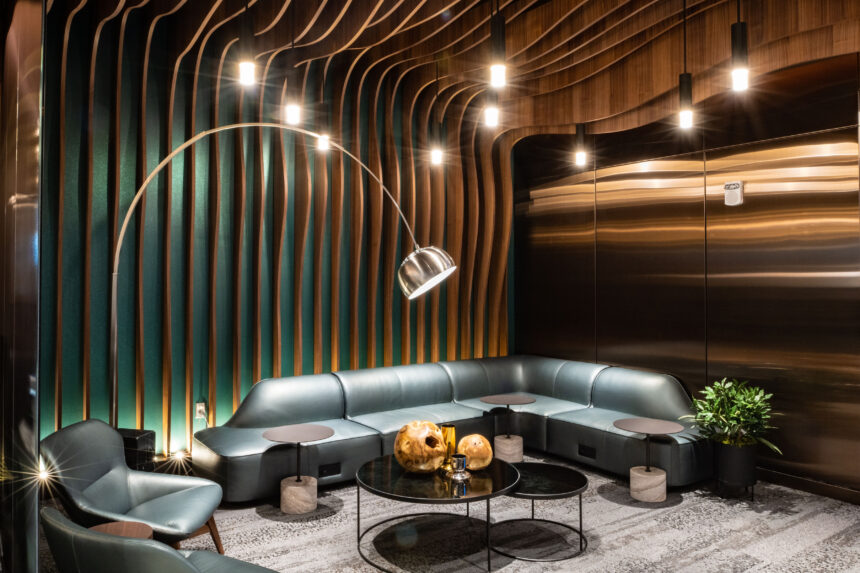 modern airport lounge interior with curved wood and teal wall paneling, soft leather seating, round black coffee tables, ambient pendant lighting, and a large arched floor lamp.