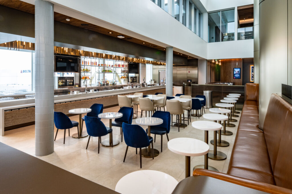 Spacious and modern Delta lounge featuring a sleek bar with backlit shelving, blue and beige lounge chairs, white marble-top tables, and floor-to-ceiling windows. The design includes high ceilings, neutral tones, and natural lighting,.