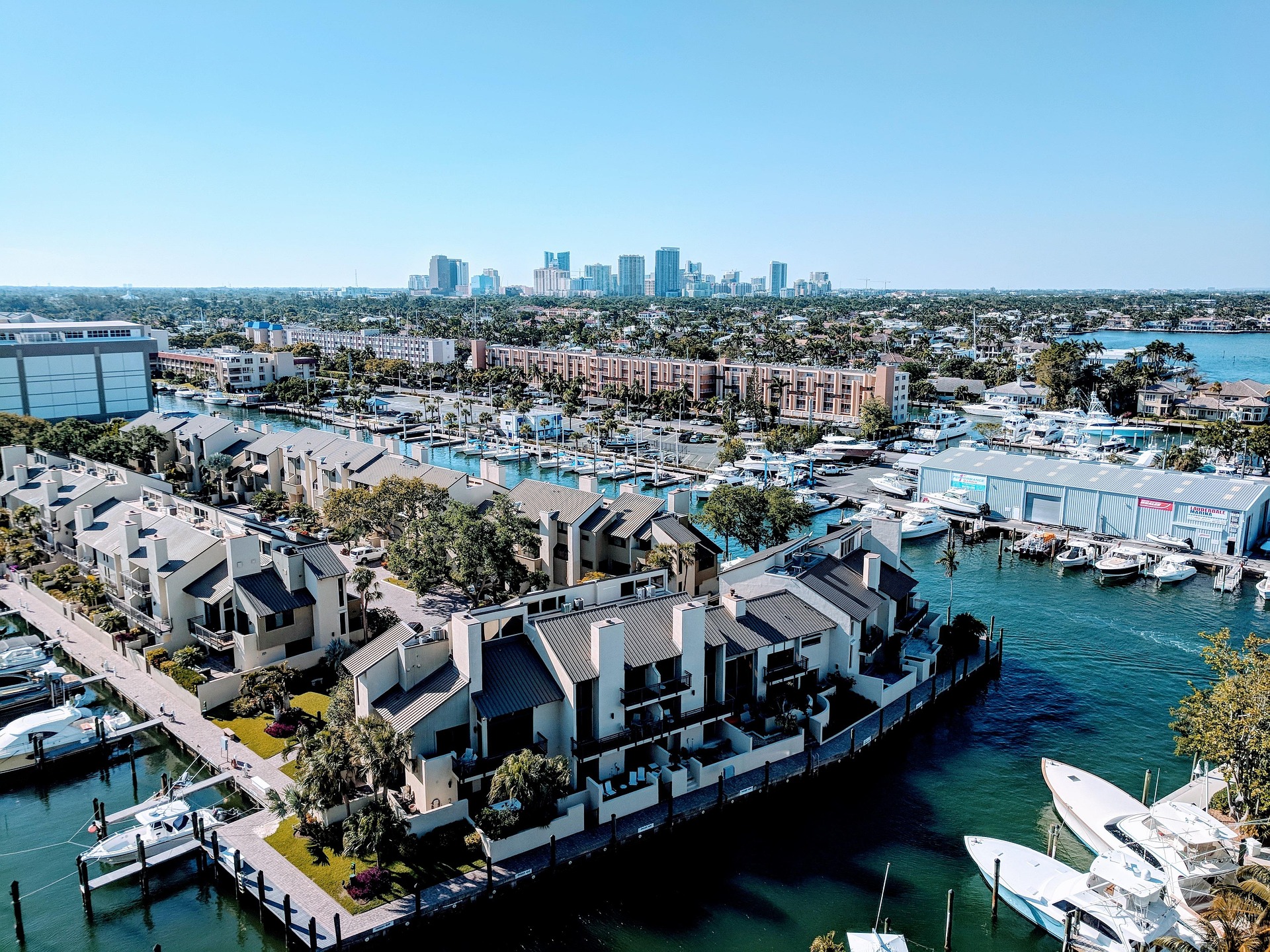 Aerial view of Fort Lauderdale, Florida, showing waterfront homes, marinas with docked boats, palm trees, and a city skyline in the distance under a clear blue sky.