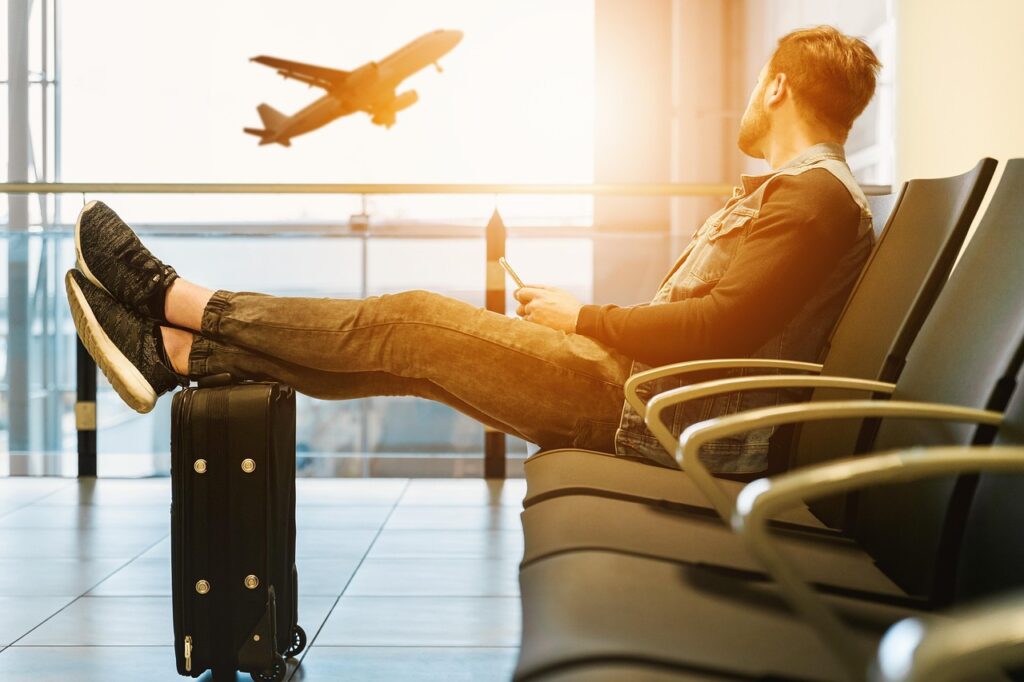 A man relaxes in an airport lounge with his feet on a suitcase, watching a plane take off through the window as sunlight streams in