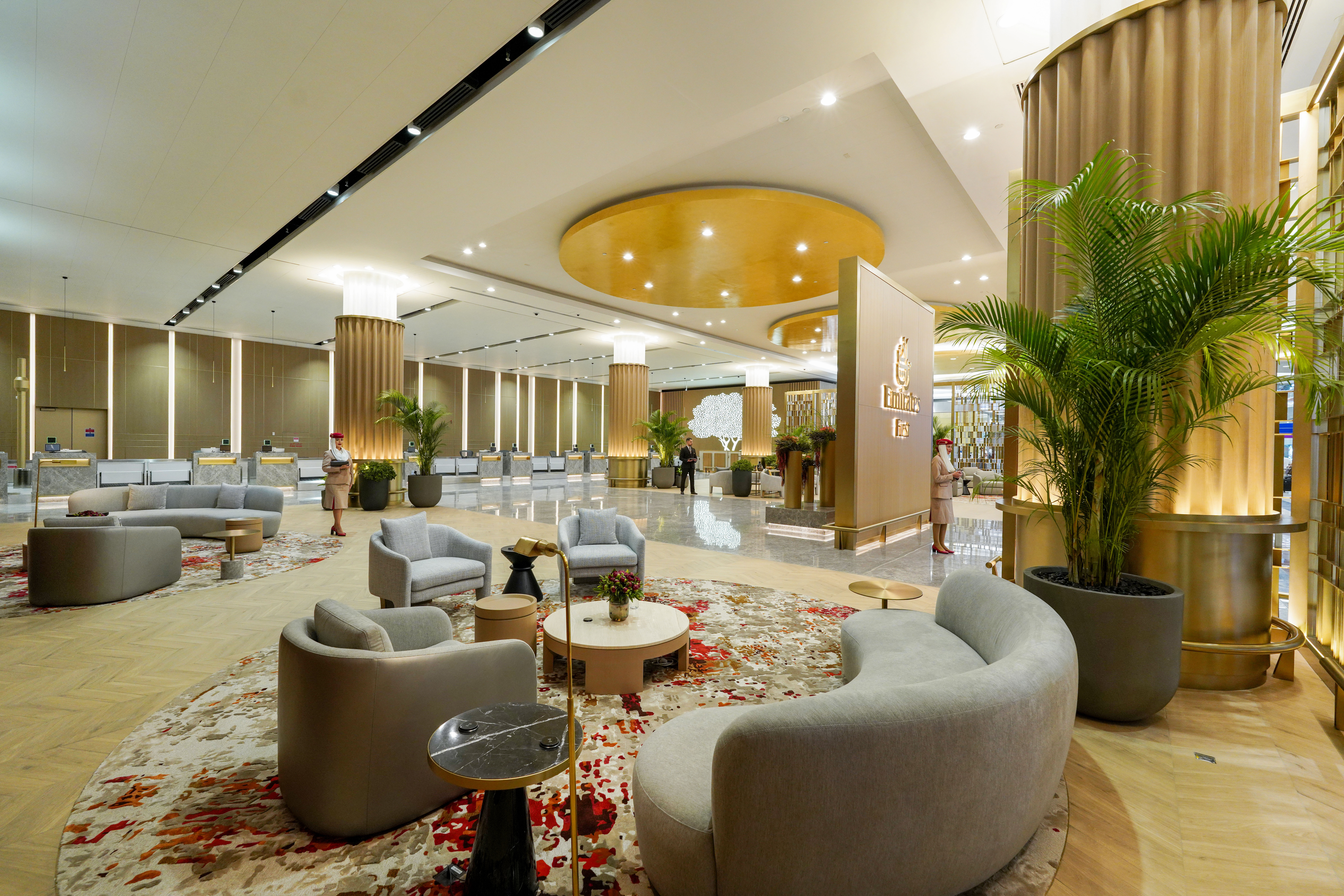 A luxurious Emirates First Class lounge featuring elegant grey and beige seating areas with red-accented rugs, gold-toned columns, lush indoor plants, and a prominent Emirates First sign. The check-in counters are visible in the background, with airline staff in uniform attending to passengers.