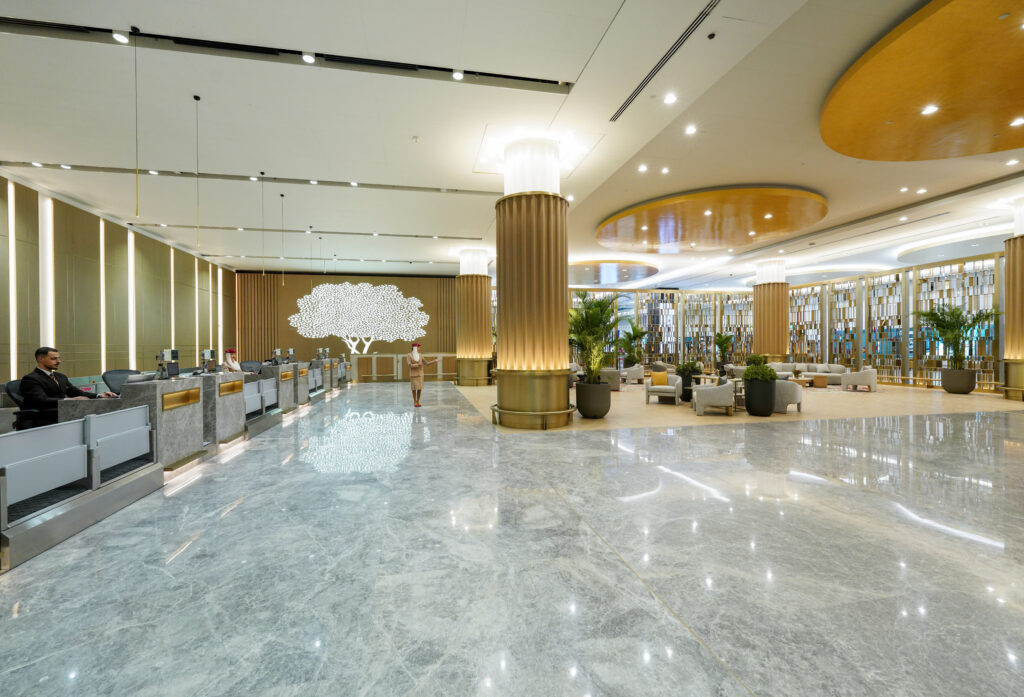 A spacious and luxurious airport lounge featuring a row of check-in counters on the left, plush seating areas on the right, elegant gold columns, marble flooring, and a large illuminated tree design on the back wall.