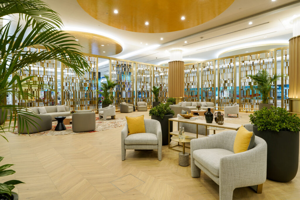 A refined seating area inside the Emirates First Class lounge at Terminal 3 at DXB, featuring modern grey and beige furniture with yellow accent cushions, decorative plants, and golden partition panels that add privacy and elegance to the open-plan layout.