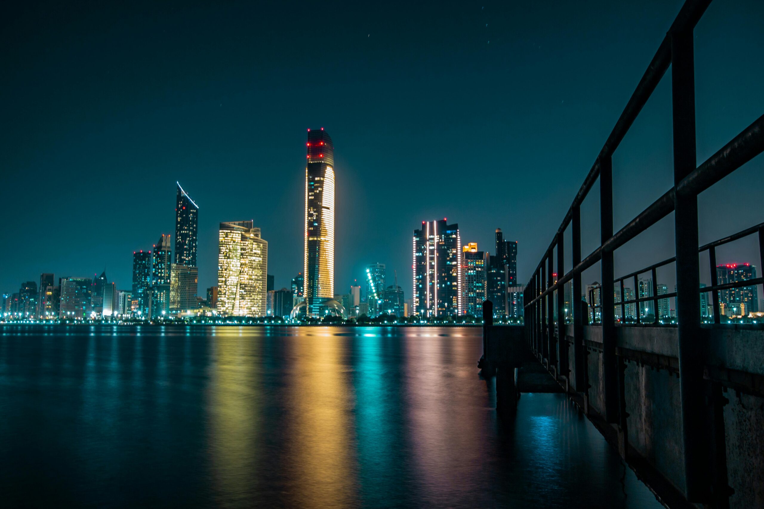 Abu Dhabi's skyline at night, with skyscrapers and city lights reflected in the water below.