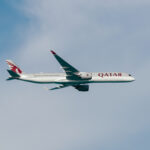 A Qatar Airways airplane is flying in the sky. The aircraft is a modern jet with a sleek design, featuring the airline's logo and branding on its fuselage and tail. The background shows a clear blue sky with some light clouds.