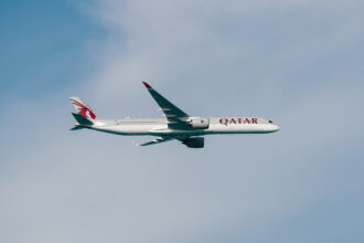 A Qatar Airways airplane is flying in the sky. The aircraft is a modern jet with a sleek design, featuring the airline's logo and branding on its fuselage and tail. The background shows a clear blue sky with some light clouds.