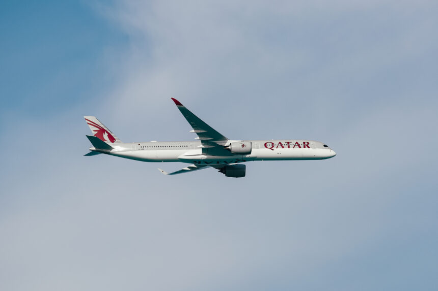 A Qatar Airways airplane is flying in the sky. The aircraft is a modern jet with a sleek design, featuring the airline's logo and branding on its fuselage and tail. The background shows a clear blue sky with some light clouds.