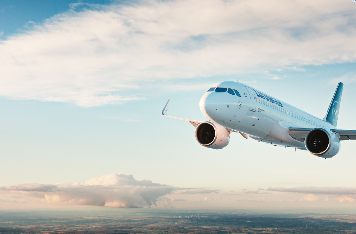 A Lufthansa airplane is flying in the sky with a backdrop of clouds and a landscape below. The aircraft is angled slightly upward, showcasing its engines and wings.