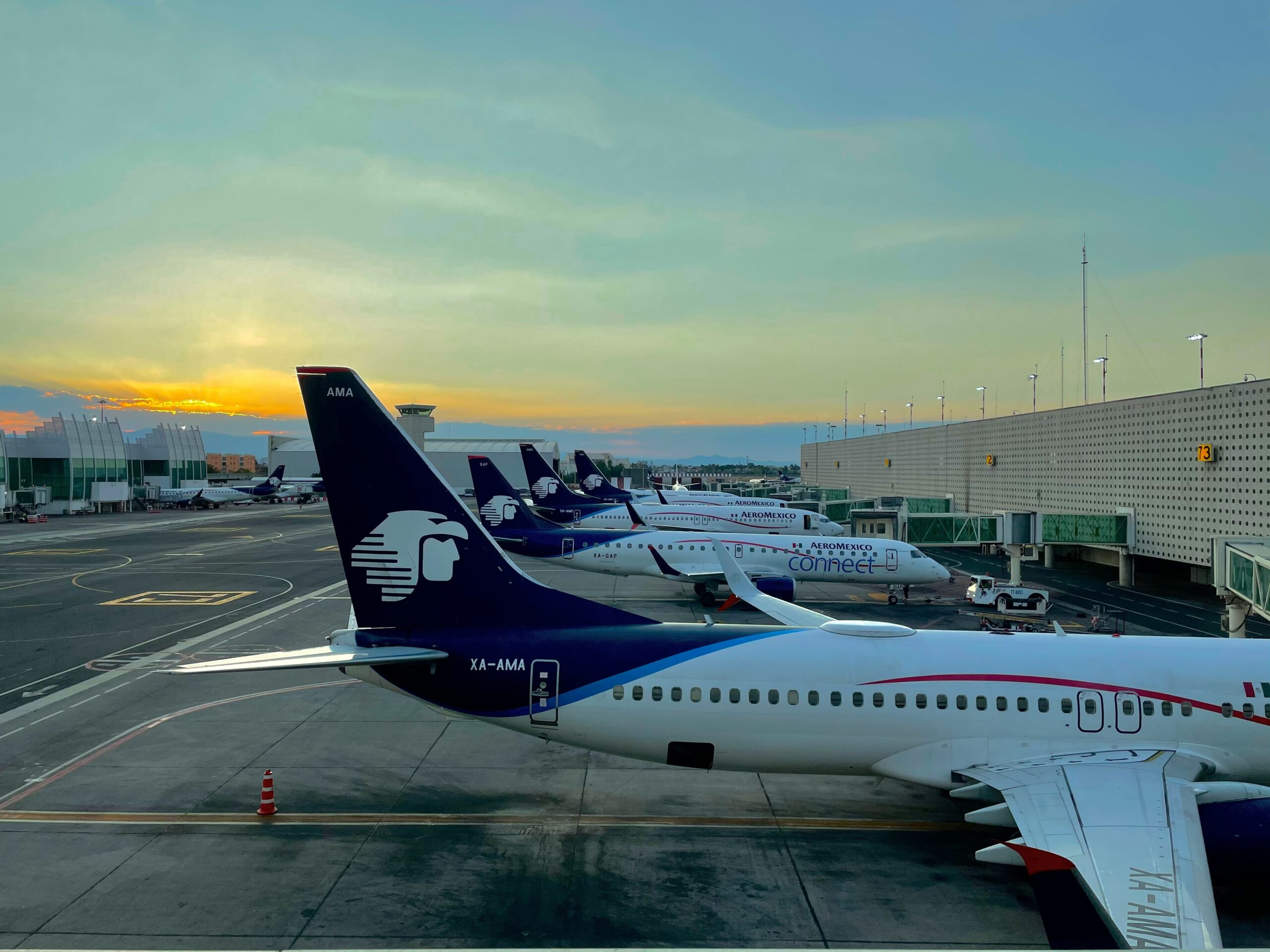A row of Aeromexico aeroplanes parked at gates in an airport during sunset. The planes are lined up with their tails prominently displaying the Aeromexico logo, which features an eagle head. The sky is a gradient of blue and orange as the sun sets behind the terminal building.