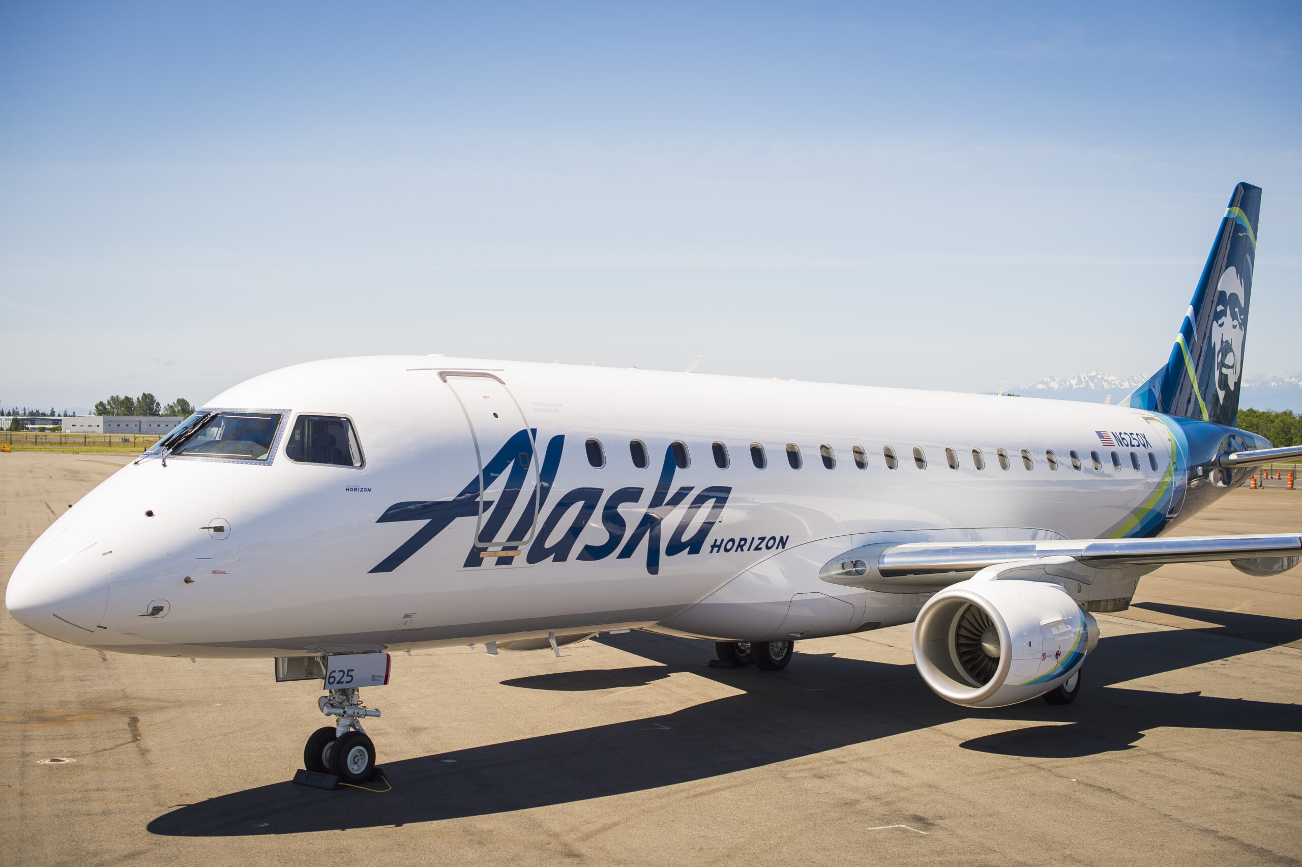 A white Alaska Horizon aeroplane is parked on the tarmac under a clear blue sky. The aircraft features the word "Alaska" prominently on its fuselage and has an image of a face on its tail fin. 