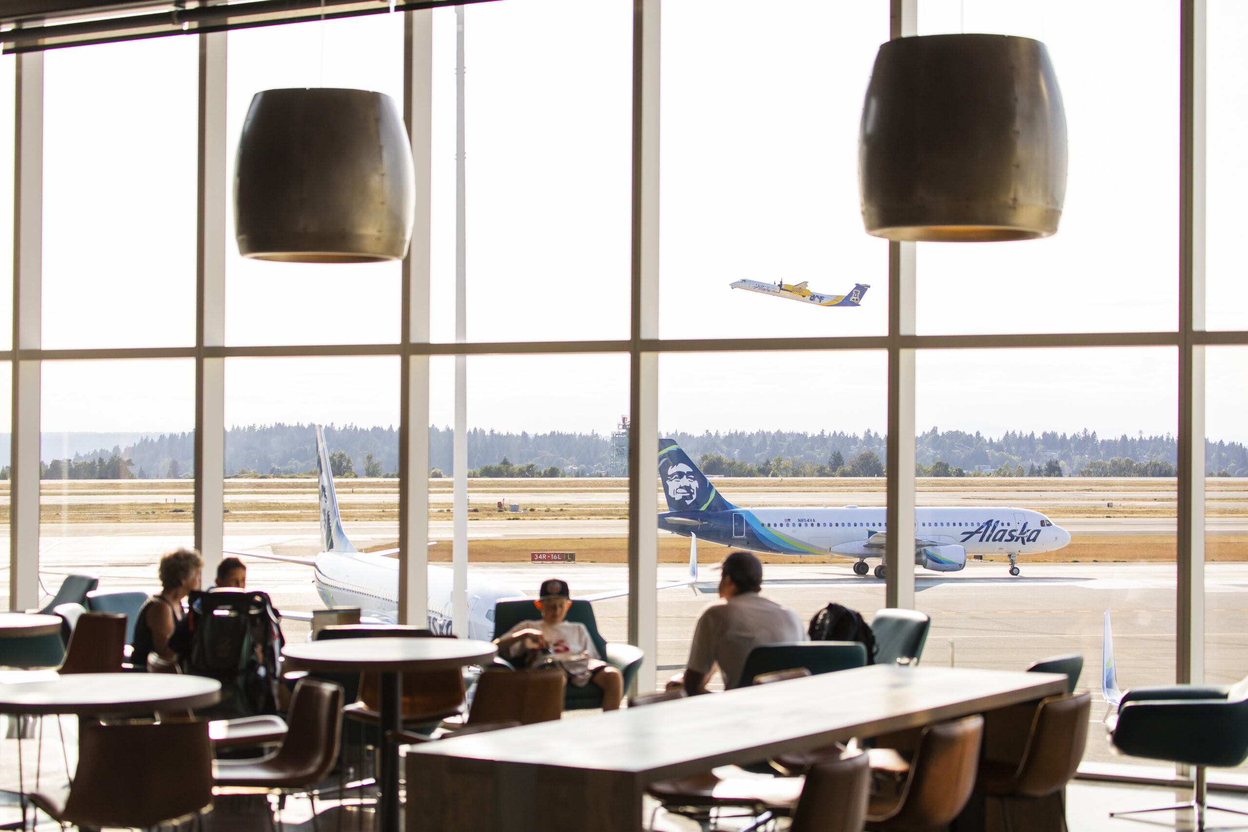 The image shows an airport terminal with large windows overlooking the runway. Inside the terminal, several people are seated at tables and chairs, some engaging in conversation. Outside on the runway, an Alaska Airlines plane is parked while another plane is taking off in the background. The scene is bright and clear with a view of trees and hills in the distance.